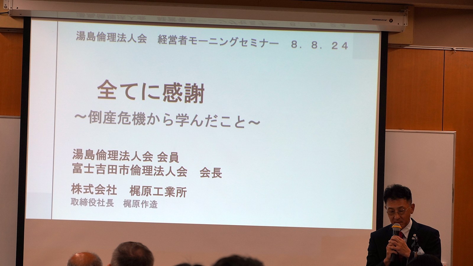 演題スライドを背に語りはじめる梶原作造氏と、聴き入る会場